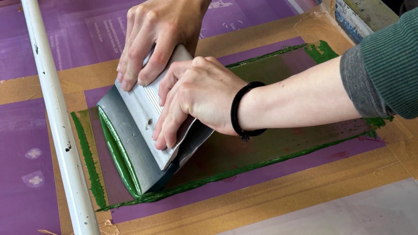 Hands pulling a squeegee across a screenprinting frame in the organiser’s workshop visual