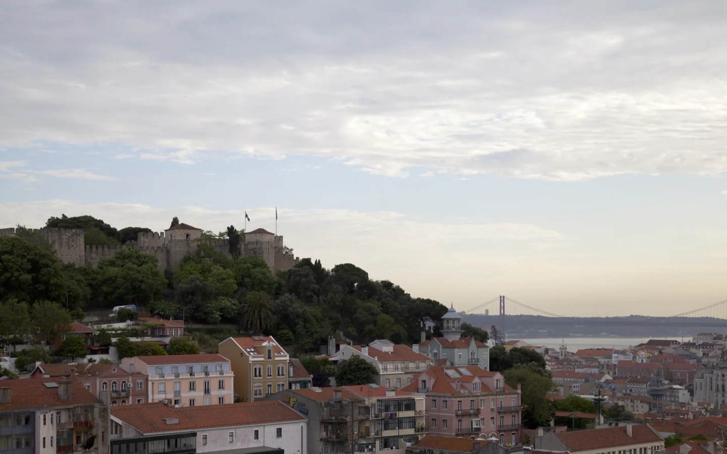 São Jorge Castle and Lisbon's red roofs seen from Miradouro da Graça, with the Tagus and 25 de Abril bridge in the distance.