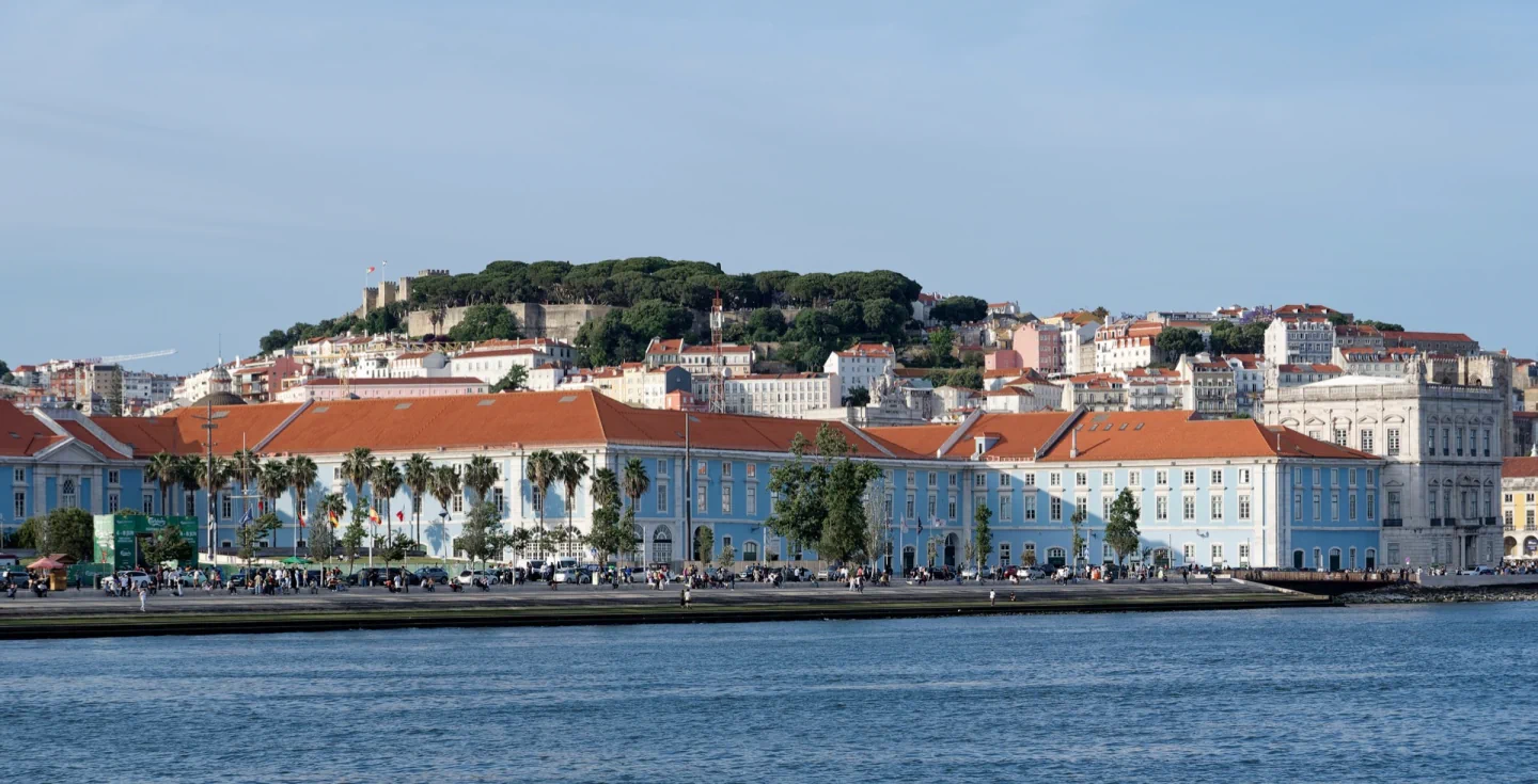 Blue waterfront buildings along Ribeira das Naus with red roofs and Castelo de São Jorge behind, seen from the Tagus.
