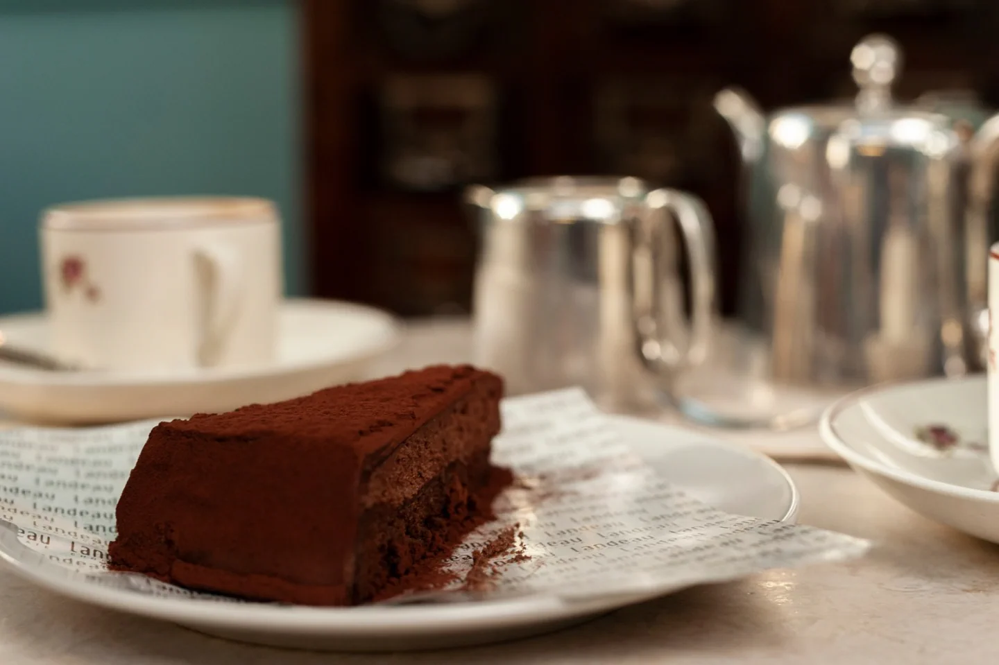 A slice of Landeau chocolate cake beside coffee and a silver teapot